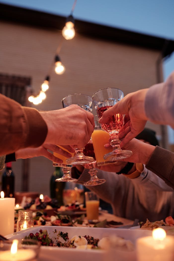 Crop friends gathering at table under burning garland in garden and toasting with glasses